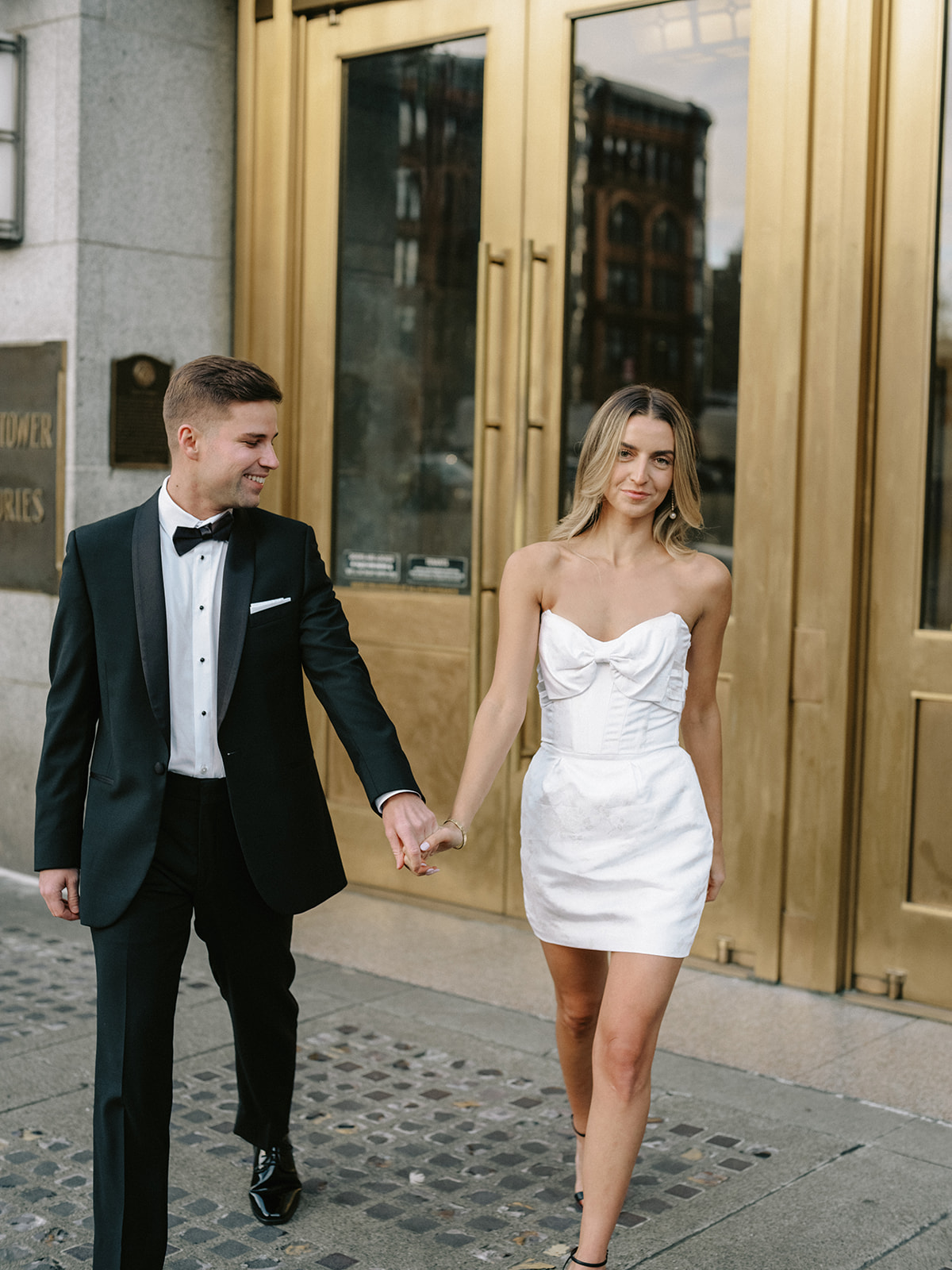 girl in white dress and boy in black suit walk away from building